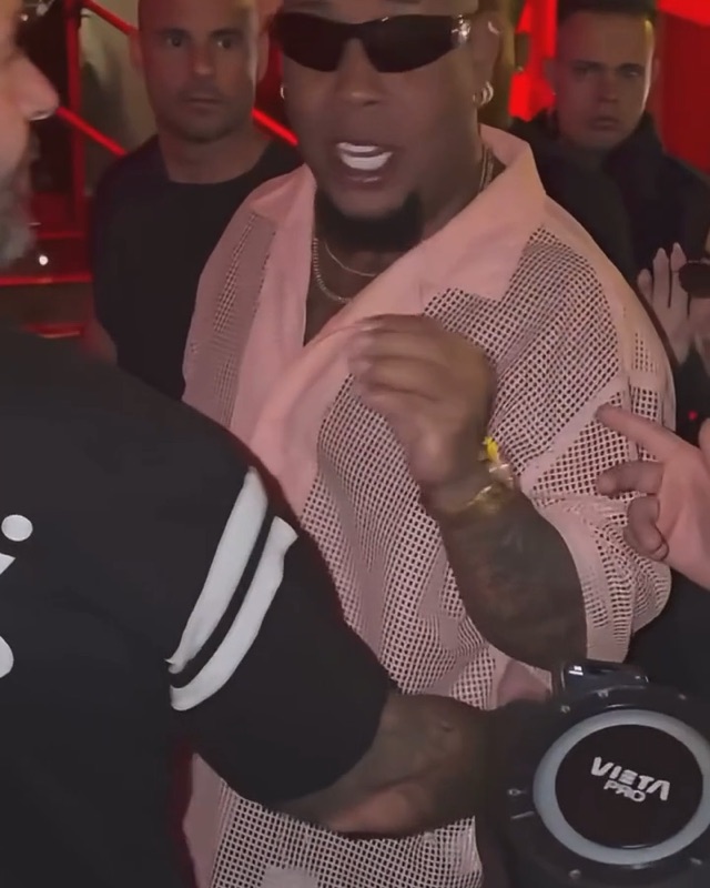 Dame Hombro AI clip opening: a fan holds a portable speaker up to a man in sunglasses and a pink mesh shirt in a red-lit entrance